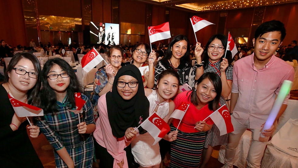 A joyful group of people holding Singapore flags in a festive indoor setting, decorated with warm lighting and a large crowd in the background.