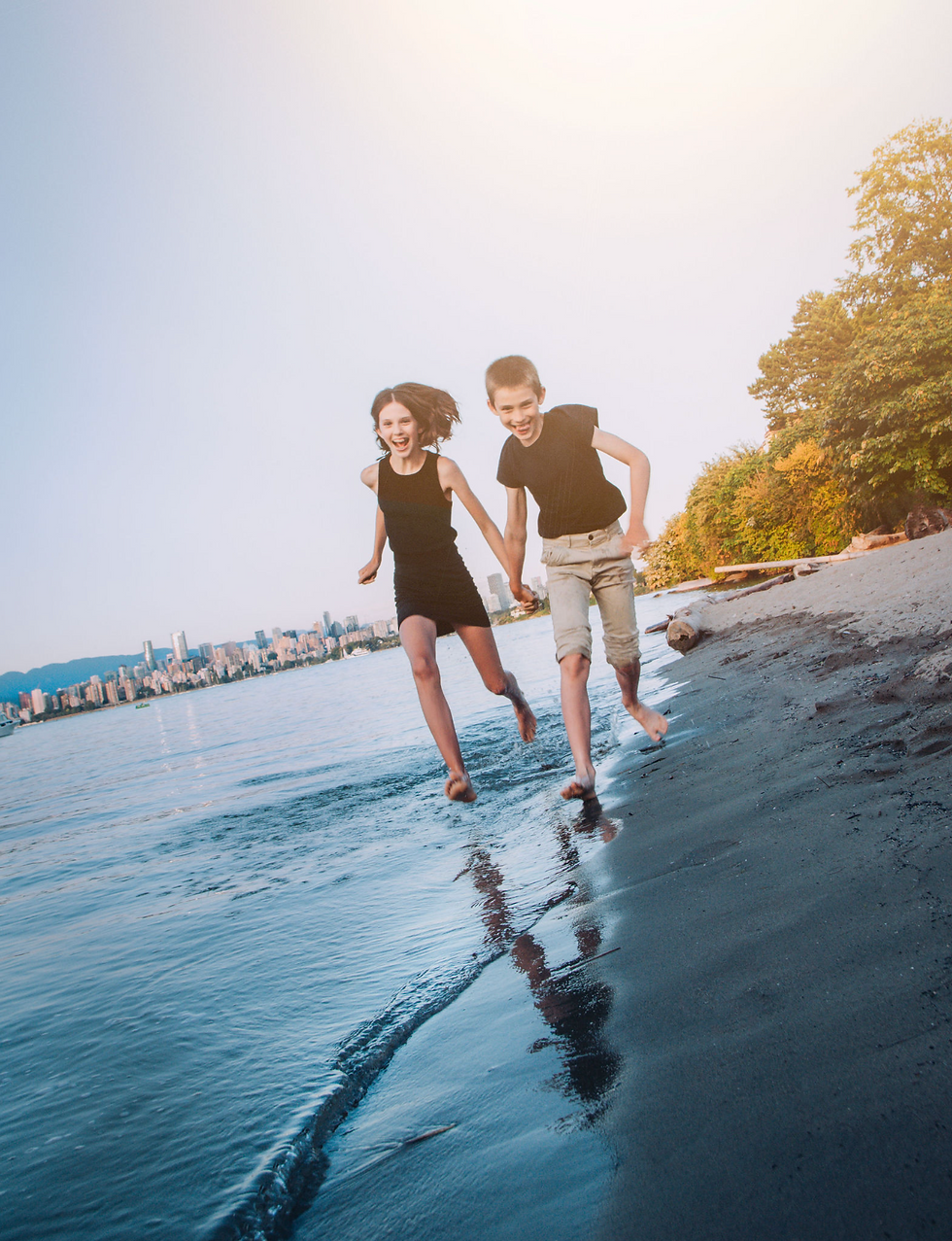 Siblings running barefoot along the shoreline at Jericho Beach in Vancouver with city skyline in the background – playful family photo by My City Photos.