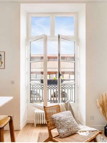 Bright room with wicker chair, art, and open window view of rooftops. Warm tones, minimal decor, cozy and serene atmosphere.