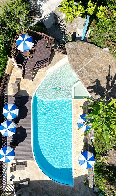 Vista aérea vertical de uma piscina aquecida com água cristalina e bordas brancas, cercada por área de deck com piso de ladrilho e madeira. Há guarda-sóis azuis e brancos distribuídos ao redor, cadeiras de sol e vegetação tropical nas proximidades, incluindo bananeiras e grandes pedras naturais.