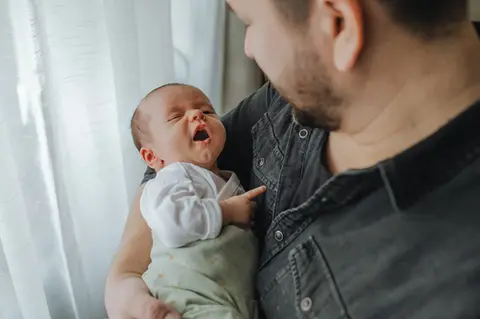 Vater hält sein Neugeborenes im sanften Fensterlicht während einer Homestory in Liesing, Wien, fotografiert von Pia Morpurgo Fotografie.