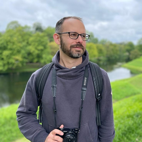 A bearded man with brown hair and glasses stands in front of a blurred grassy background. A camera hangs around his neck.