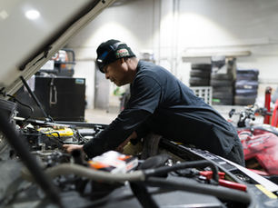 Mechanic in a Castrol EDGE beanie leans into an open car engine in a garage, focused under bright work lights.