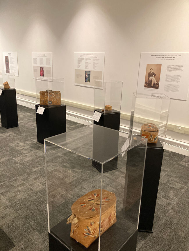 4 birchbark baskets in exhibition cases for To Honour and Respect exhibit. Text panels with images on wall behind the cases.