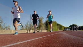 Three runners jog on a red track under a clear blue sky, with a chain-link fence and trees beside them.