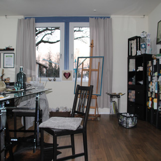 ein Stuhl steht an einemTisch, ein Regal mit Wolle steht rechts an der Wand, im Hintergrund sieht man ein Fenster, davor steht ein Bild mit einem Herz