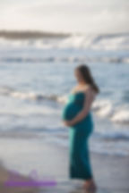 Pregnant woman in turquoise dress holds her belly while looking down at the sand during a photo shoot on a Maui beach
