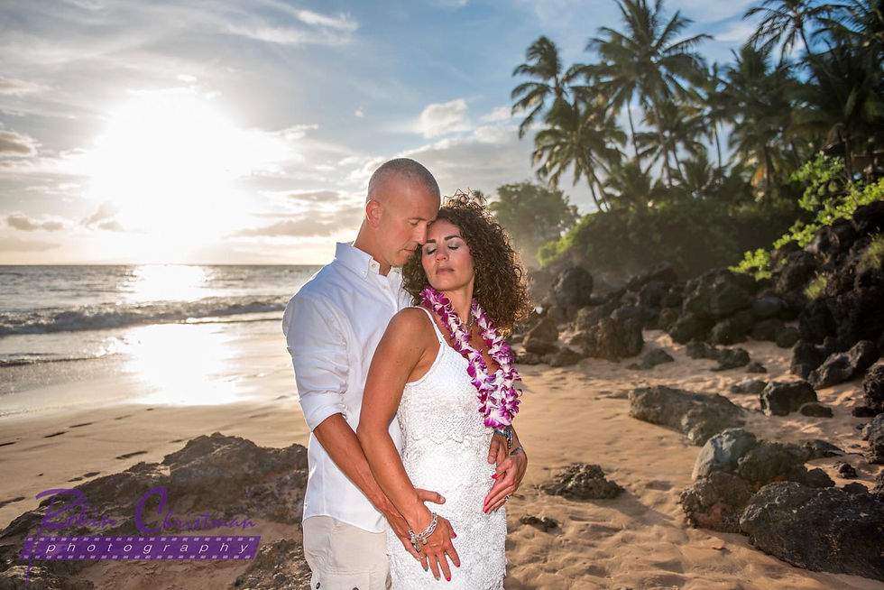 Man embraces wife from behind with eyes closes as they stand on the shoreline of a beach on maui