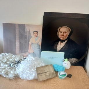A table containing two polyester lawyers' wigs, a pair of glasses, a grimy mid-eighteenth-century will folded into a square, clay, a small pile of seventeenth and eighteenth century coins, blue cards with questions on, a small ball of turquoise wool, a white squishy carrot, a poster of an eighteenth century painting of a young Black edinburgh milk seller, and a poster of a seventeenth century painting of an older woman in dark clothing and cap