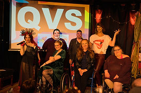 A photo of the cast of the Queer Variety Show in August 2024. featuring, Bambi Valentine, Cody Smith, Sparklemuffin, Holly Hazlewood, Sweet Az, Ginger Nibbles and Temerity Outře. Behind them is a screen with the Queer Variety Show logo. They are all smiling. Photo by Ian Chua.