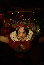 Bambi Valentine is dressed as the Queen of Hearts. Whe has dark hair and eyes, and is a white skinned person. She is wearing a headdress made of roses and has a red collar, with playing cards as a part of it. Her image disappears into the dark background. the background is dark with fairy lights. She is staring down the camera.