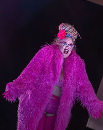 Dread sexy stands against a black backdrop, she is a light skinned woman with her blonde and pink dreads piled in a massive pile on top of her head with a pink flower. She is wearing a bright pink fluffy jacket, bra, undies and stockings. She has heavy makeup on her face and wearing sparkly pink glasses, looking away from the camera with her arms stretched out. She has an excited expression on her face.