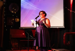 Nimona Nova is caucasian with dark purple hair. Here they are standing on a stage in front of a projection screen, reading from a sheet of paper while next to a standing microphone. They are wearing glasses, a sleeveless purple dress with a belt, and socks with black and purple horizontal stripes.