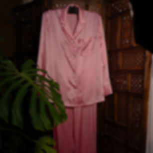 Pink satin silk pajama set with button-down top and pants, hanging on a carved wooden screen.