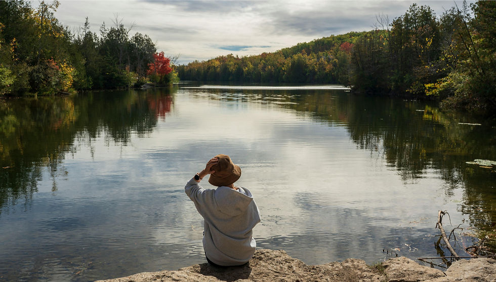 Person in a hat sits by a calm lake surrounded by autumn trees. The sky is overcast, reflecting on the water. Peaceful and serene mood.