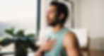 Man in a green tank top sits indoors, eyes closed, hand on chest, appearing calm. A plant and bright window are in the background.