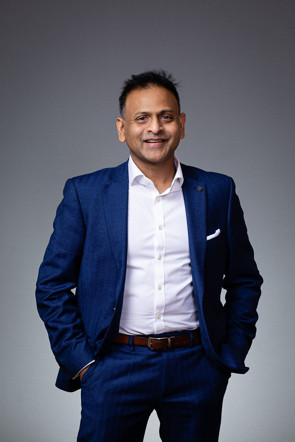 Smiling man in a navy suit and white shirt against a gray background, hands in pockets, exuding confidence.
