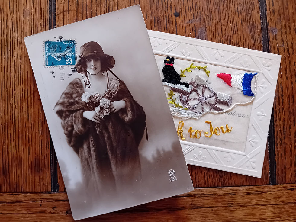 Two postcards lie on a wooden surface. On the left, an elegantly-dressed woman looks out at the viewer, holding a bunch of flowers. On the right, an embroidered postcard shows a cannon, French flag and a black cat.