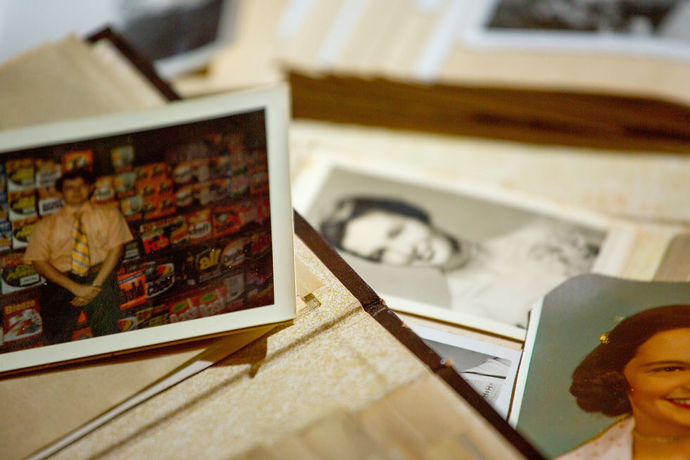 Close-up of vintage photos scattered on a table. A faded color photo shows a person in front of colorful product boxes. Mood: nostalgic. Remembering the Past.