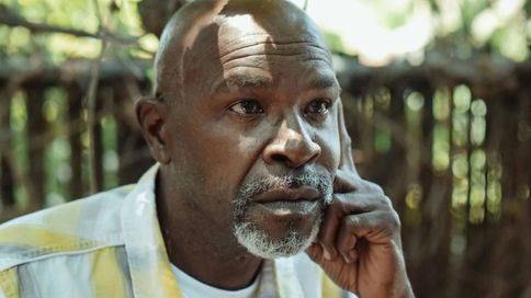 A thoughtful middle-aged Black man with a shaved head and graying beard, resting his chin on his hand while gazing into the distance in an outdoor setting.