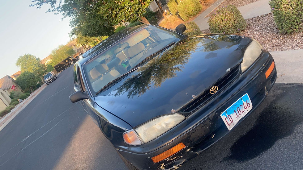 A black car with Illinois license plate parked on a suburban street. Trees and houses line the background in soft sunlight.