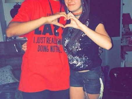 A smiling couple forms a heart with their hands. The man wears a red shirt with black text. Cozy room setting with framed photos in background.