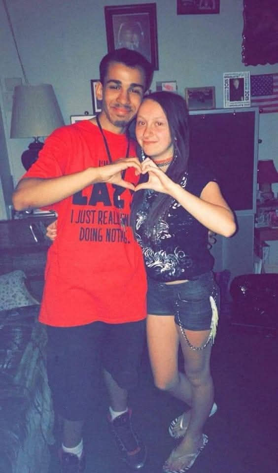 A smiling couple forms a heart with their hands. The man wears a red shirt with black text. Cozy room setting with framed photos in background.