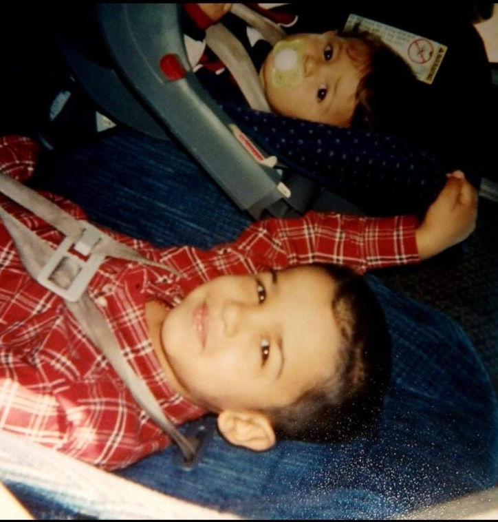 Two young children in car seats, one in a red plaid shirt smiling, the other with a pacifier. Blue fabric seats, calm mood.