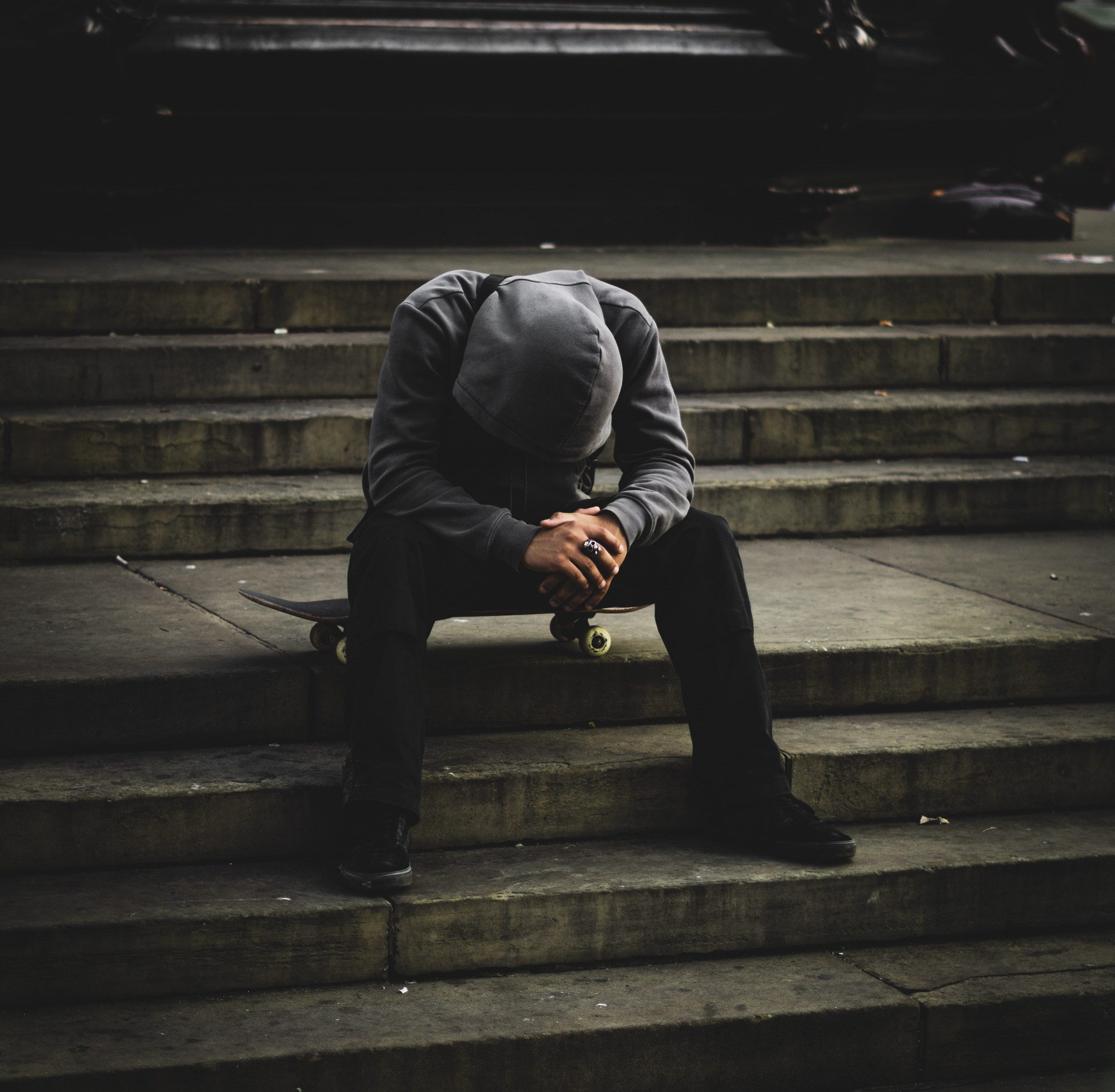 Dreary background with a person sitting on concrete outdoor steps, on a skateboard, wearing a gray hoodie with hood on, head looking down into lap with hands together