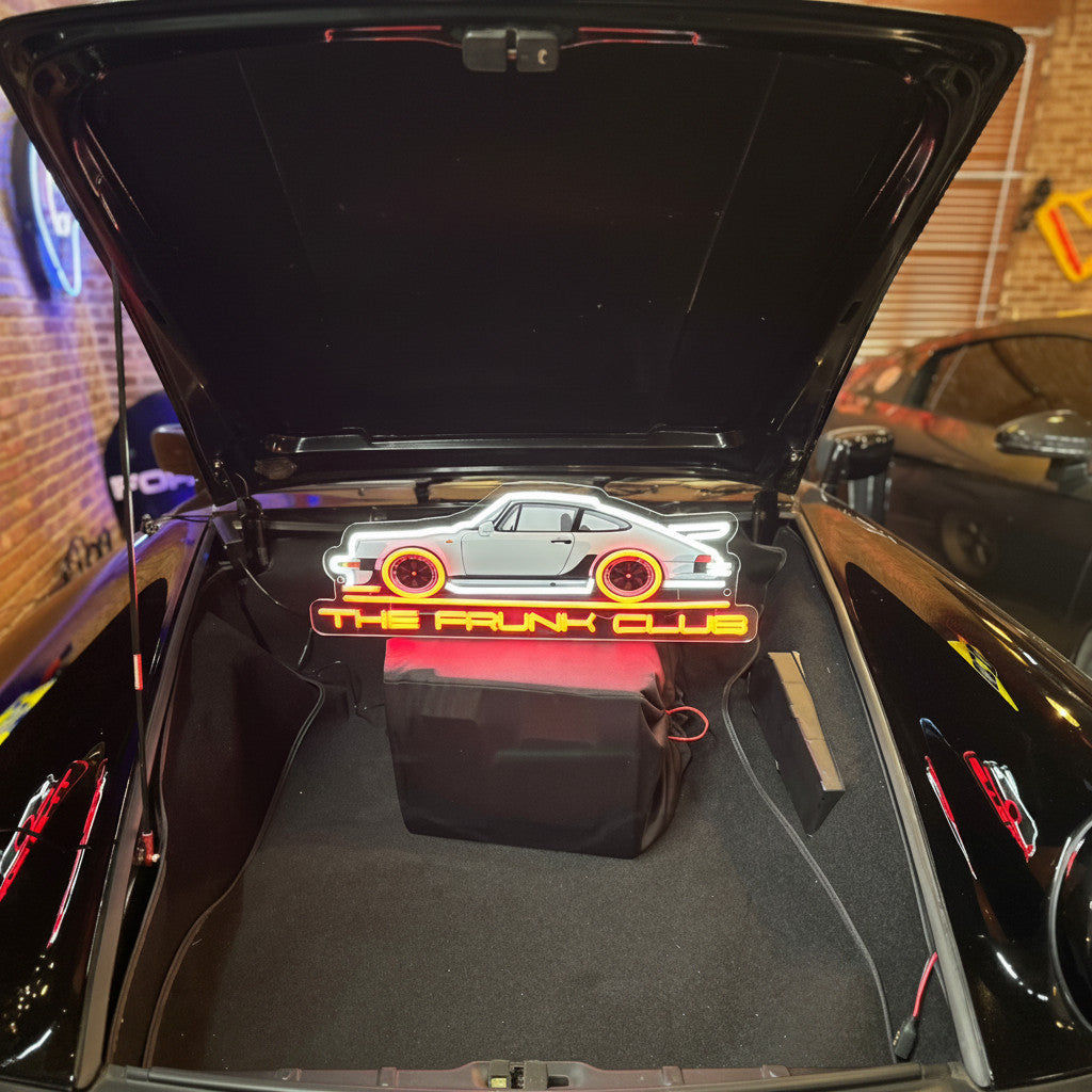 Open front trunk of a sports car with a rectangular neon sign box displaying a stylized car outline and “THE FRUNK CLUB.”