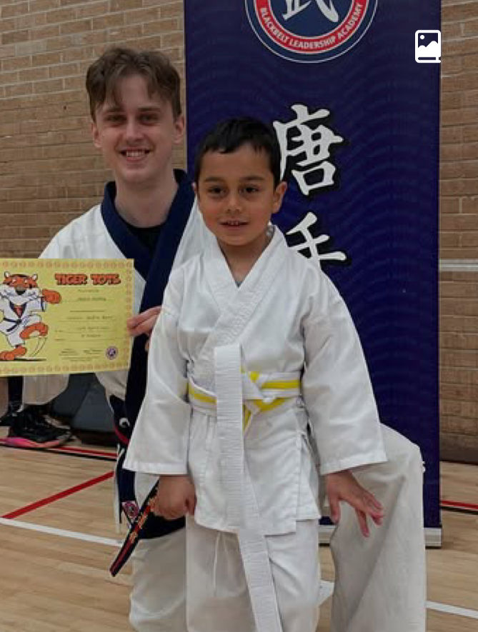 Two boys in martial arts gis, one holding a Tiger Tot certificate, posing in a gym before a blue banner.