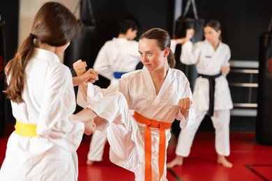 Two women in white karate uniforms sparring in a do jang with red mats. One wears an orange belt, the other yellow. Focused and intense mood.