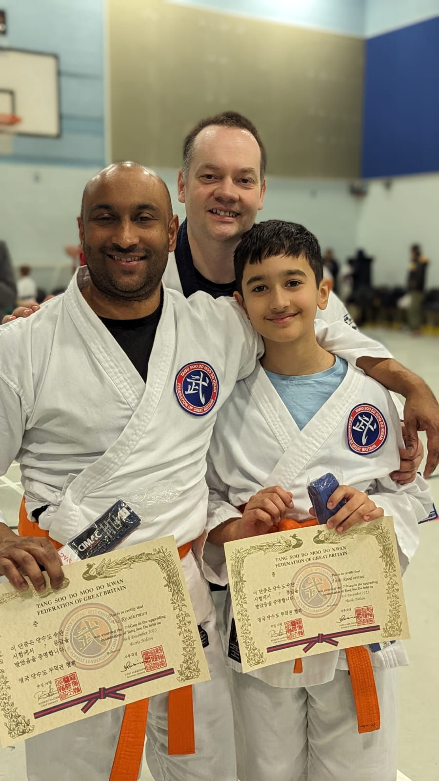 Three martial arts students in white gis and orange belts smile in a gym, holding certificates after a grading ceremony.