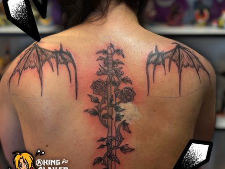 Black and grey back tattoo featuring a sword running down the spine with roses and vines growing around the blade, framed by large tattered wings symbolizing strength and sacrifice.