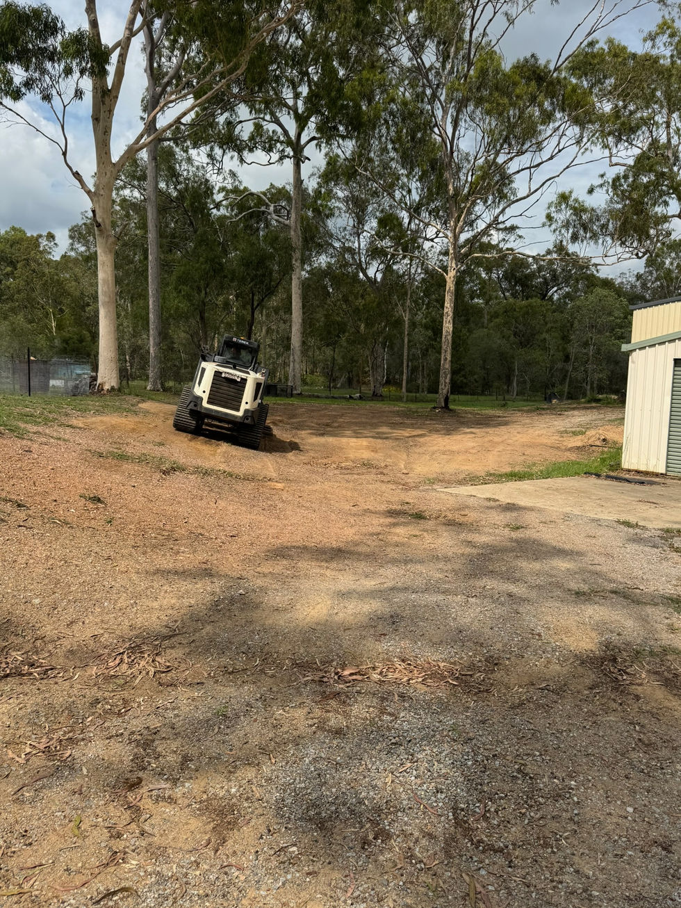 Skid steer on dirt site with trees and a building