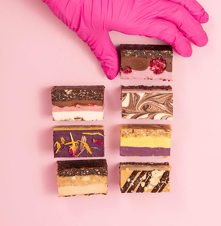 Ein Flatlay von verschiedenen angeschnittenen veganen glutenfreien Törtchen auf rosa Grund, mit einer danach greifenden Hand.