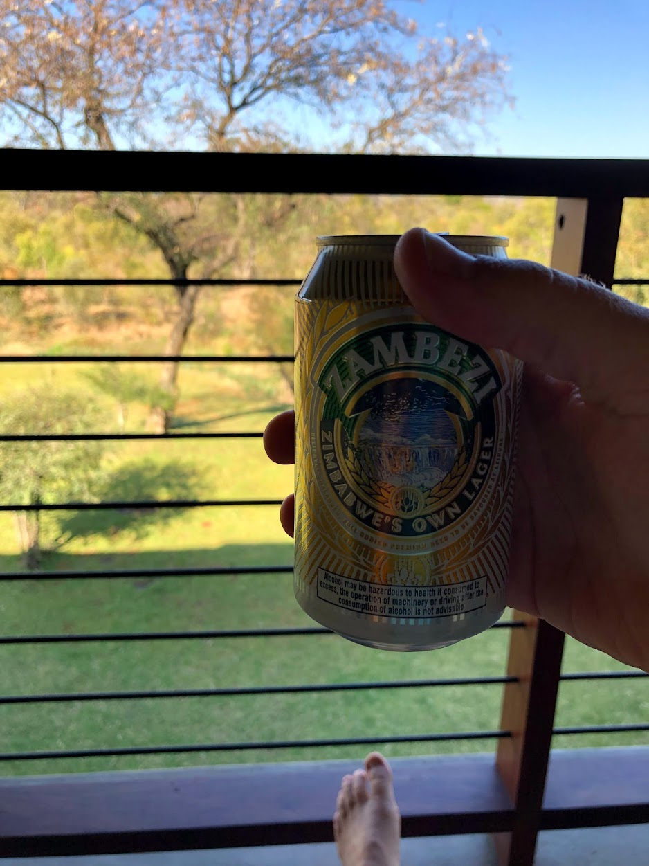 Hand holding a can of Zambezi Lager on a veranda at Ilala Lodge in Victoria Falls, with a view of green lawn, trees, and the bush landscape beyond; a bare foot is visible resting on the deck railing.