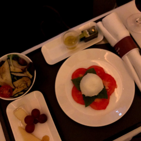 Business class meal on a Swiss Air flight, including a plate of sliced tomatoes and mozzarella, a bowl of grilled vegetables, a cheese and fruit selection, olive oil, and a glass of red wine, served neatly on a tray with a cloth napkin.