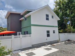 Exterior of a two-story Bon Suites Cayman building with green and white walls, a white lattice fence, and gravel parking spots labeled "PARROT" and "COCONUT."