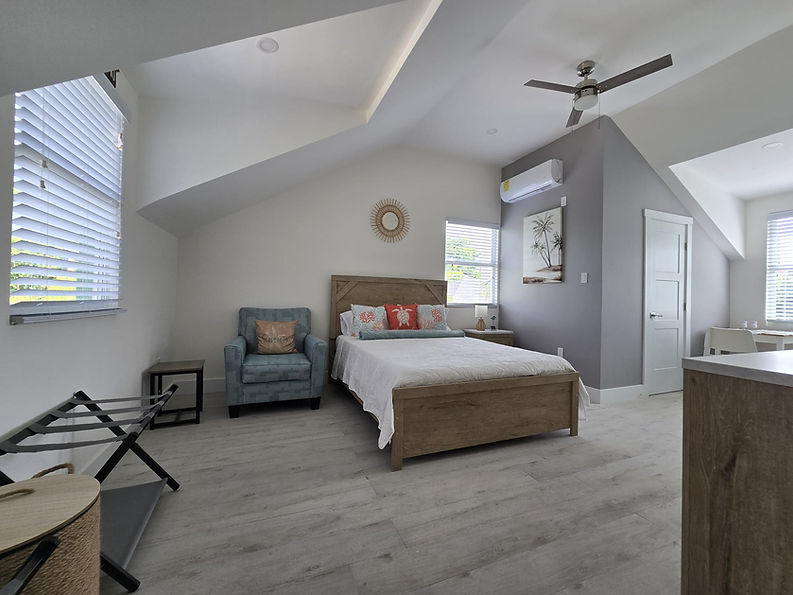 Interior of a Bon Suites Cayman studio apartment with a bed, teal armchair, white walls, and a ceiling fan.