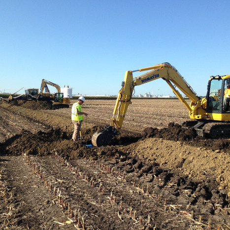 A man in a yellow vest standing next to an excavator digging a trench in a harvested field