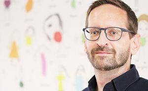 Porträt eines lächelnden Mannes mit Brille vor einer Wand mit Zeichnungen.