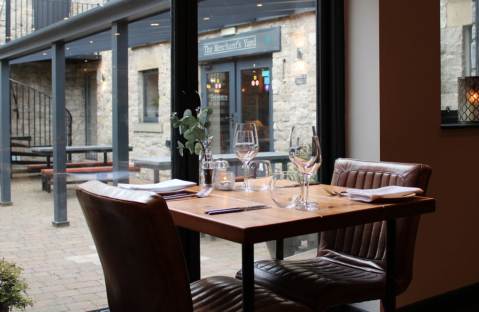Empty restaurant table set with wine glasses and plates by a window facing The Merchant's Yard courtyard.