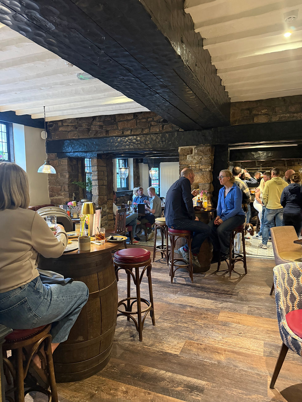 Cosy stone pub with patrons chatting at tables and bar, warm lights, wood floors, and a dog near the back.