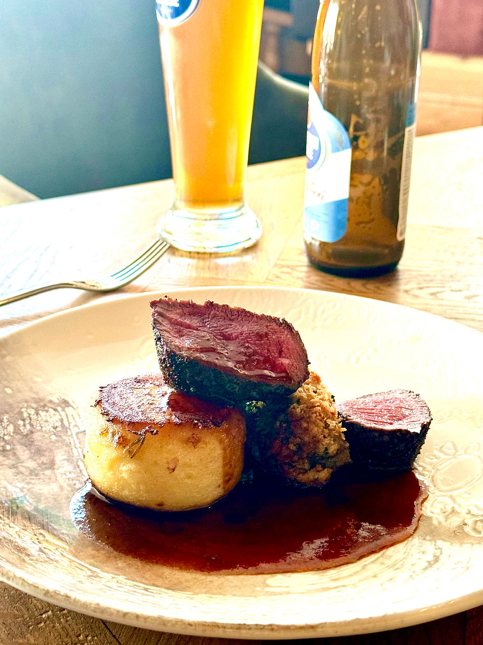 Plated steak with potato and rich sauce, beside a beer bottle and tall glass on a sunlit restaurant table.