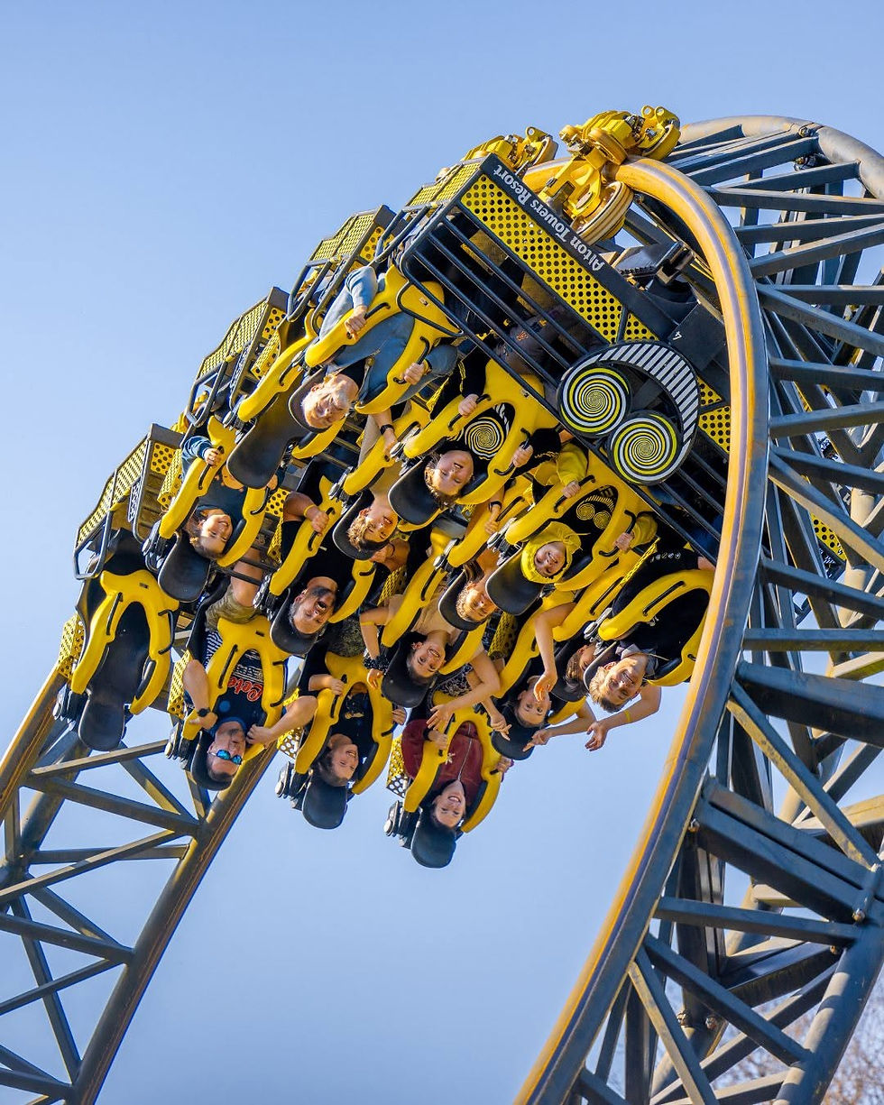 Upside-down riders on a yellow roller coaster at Afton Towers Resort against a clear blue sky, laughing.