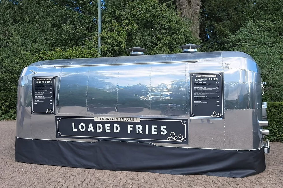 Shiny silver Loaded Fries food trailer in a park, with black menu signs and trees reflected on its polished surface.