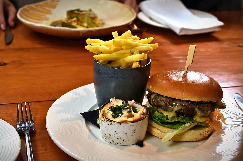Burger, fries in black cup, and slaw on a white plate at a restaurant table with warm wood tones.