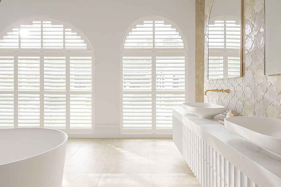 Bright white luxury bathroom with white plantation shutters, freestanding tub, double vessel sinks, arched shuttered windows, and marble mosaic wall.
