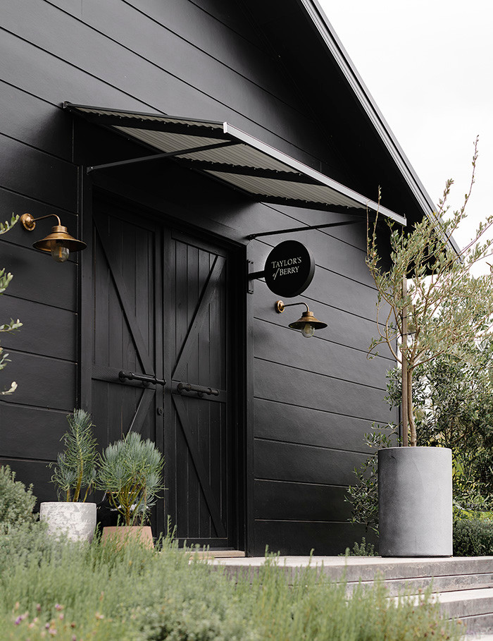 Black storefront with barn-style door, Taylor’s of Berry sign, brass lamps, potted trees, and lush garden in front.
